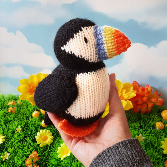 a knitted puffin being held in a hand with flowers in the background