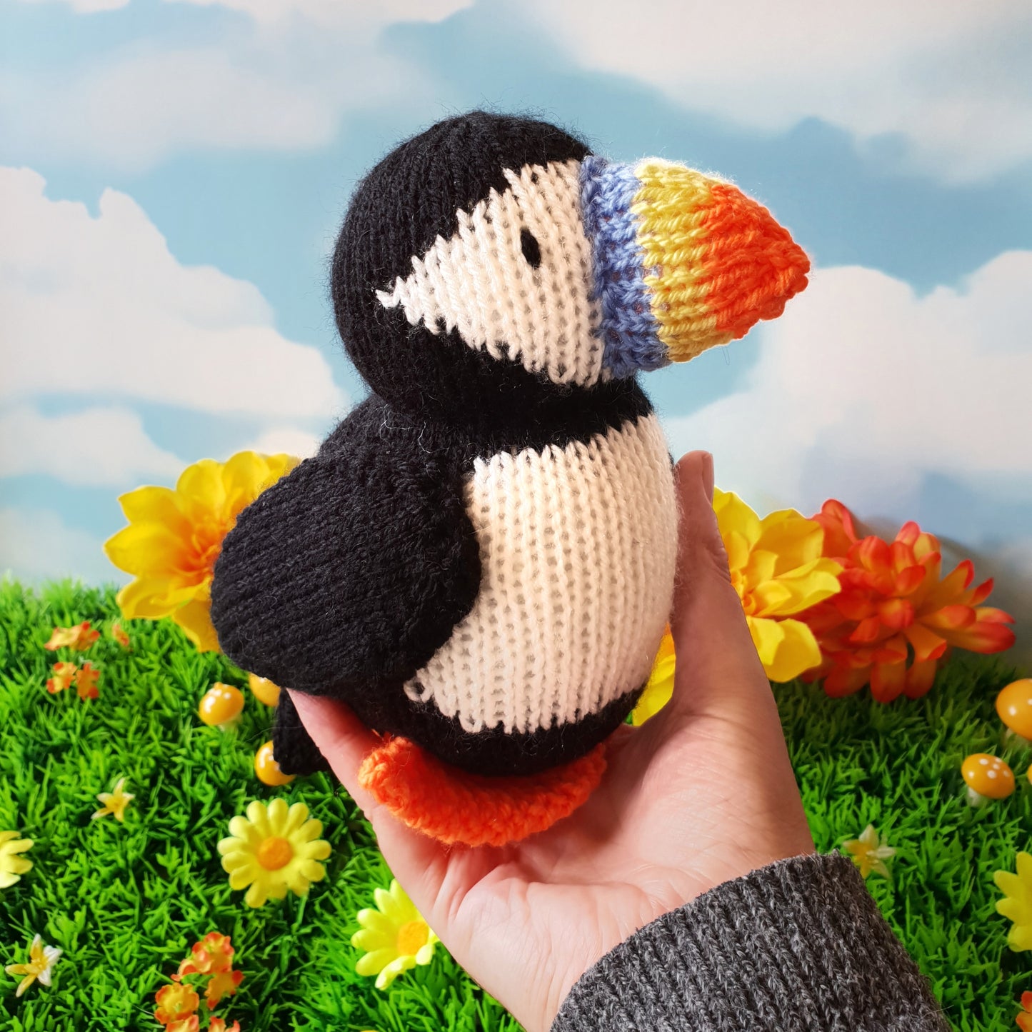 a knitted puffin being held in a hand with flowers in the background
