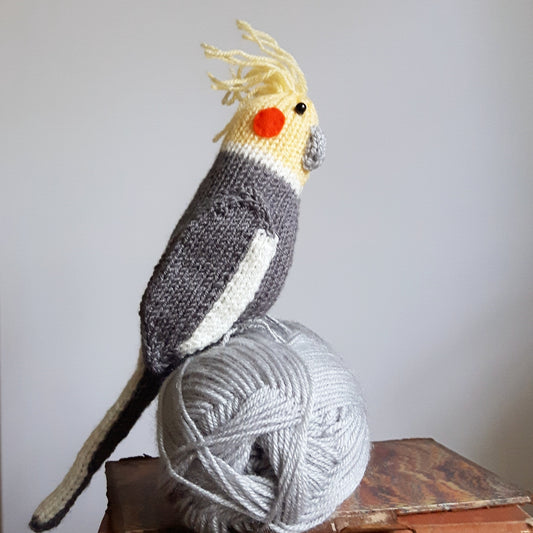a knitted grey and yellow cockatiel sitting on a ball of grey yarn