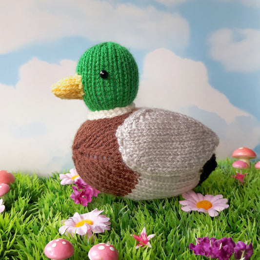 a knitted mallard duck sitting on grass with pink flowers and mushrooms