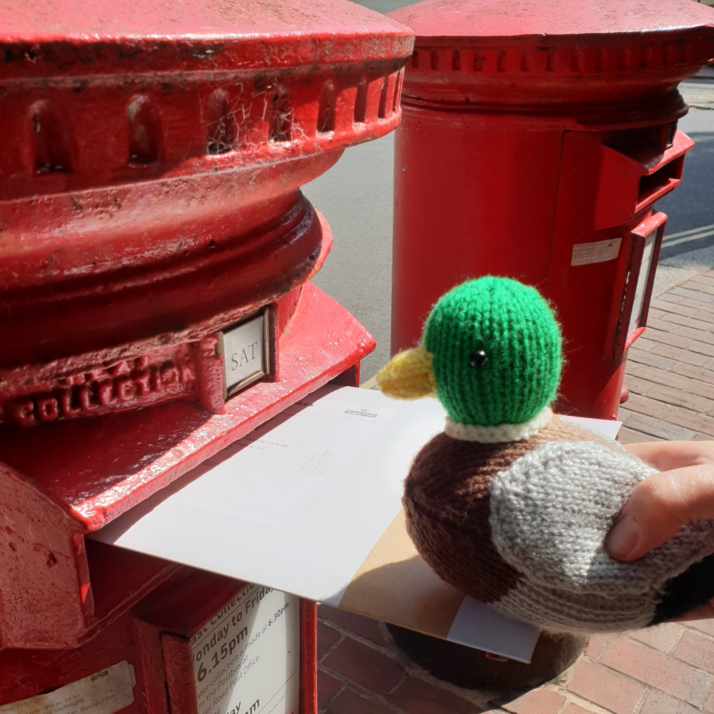 a knitted mallard duck is posting a letter in an old fashioned red postbox