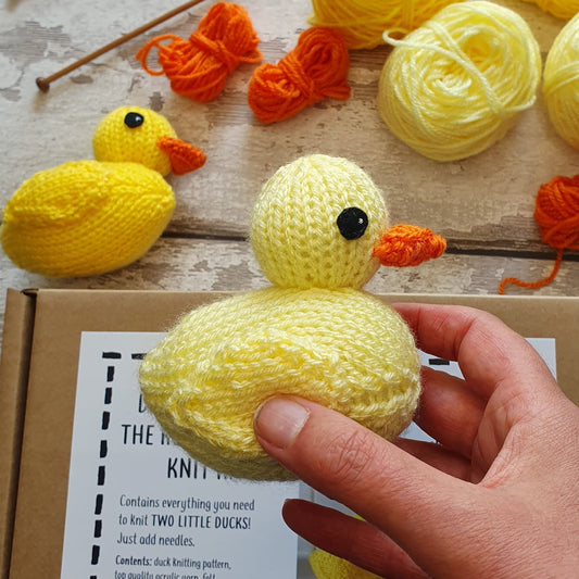 A yellow knitted rubber duck being held in a hand. In the background there is yellow and orange yarn, another rubber duck and a Nicky Stewart bird knitting kit.