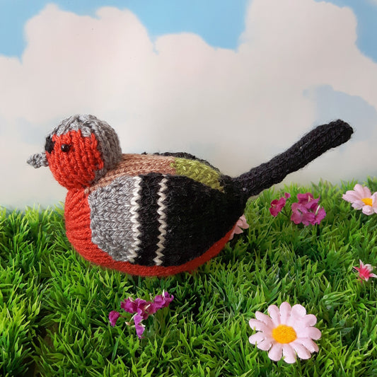 a knitted chaffinch sitting on grass with blue sky and clouds behind him