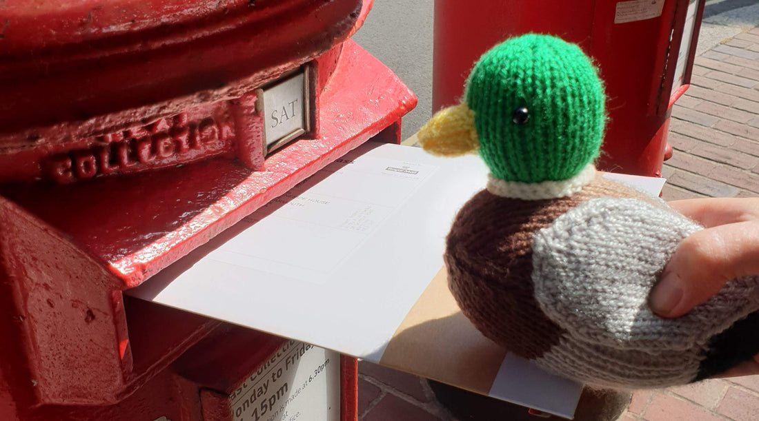 a knitted mallard duck posting a letter in a big red letter box