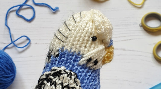 a blue and white knitted budgie on a white background with blue knitting yarn and yellow scissors in the background