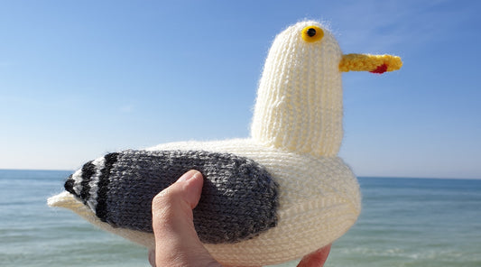 a knitted seagull being held in a hand, with a blue sea and blue sky in the background