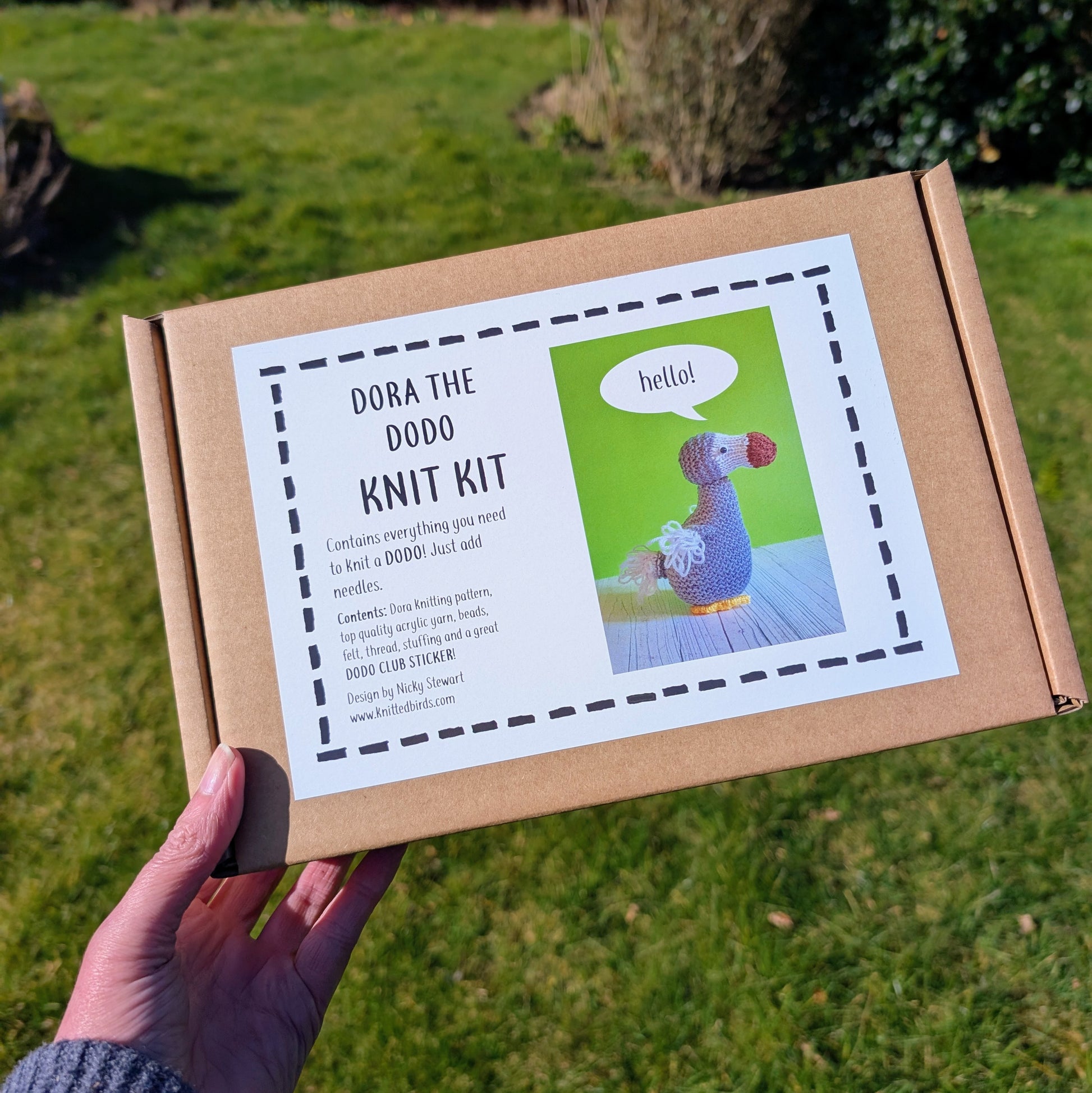 A dodo knitting kit in a gift box being held up in a garden. There is grass and hedges in the background.